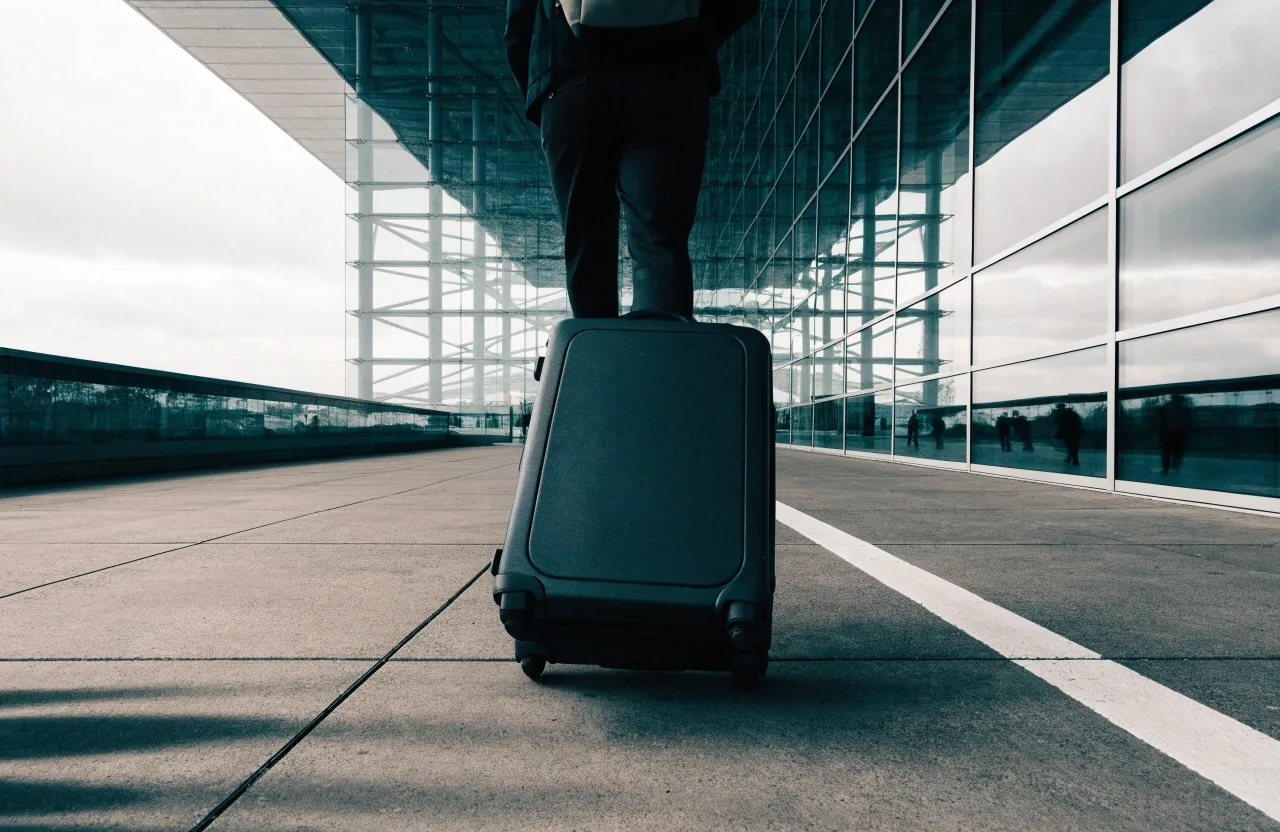 A traveller's roll-aboard suitcase seen from behind walking a marked pavement line toward a terminal building